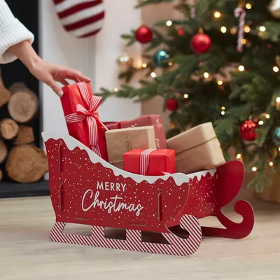 A person is placing a gift into a Christmas-themed sleigh filled with presents, with a decorated Christmas tree in the background.