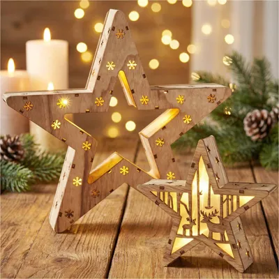 Two wooden LED star decorations are displayed on a wooden surface, with candles and pine branches in the background, creating a festive Christmas atmosphere.