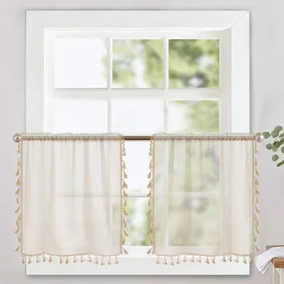 Two sheer cream-colored cafe curtains with tassels hanging on a window.