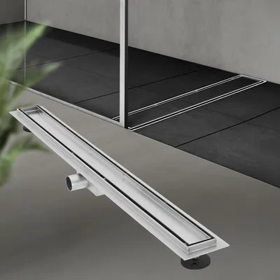 A linear shower drain is shown in the foreground, with a modern bathroom interior in the background.