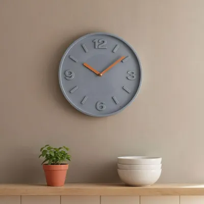 A gray wall clock with copper hands hangs on a tan wall above a wooden shelf. On the shelf, there is a small potted plant and a stack of white bowls.