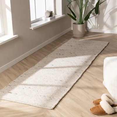 A long, narrow rug is laid out on a wooden floor in front of a window. A potted plant is visible near the window, and a pair of slippers are placed on the rug.