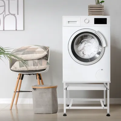 A white washing machine is shown on a white stand in a laundry room setting, next to a chair with pillows and a laundry basket.