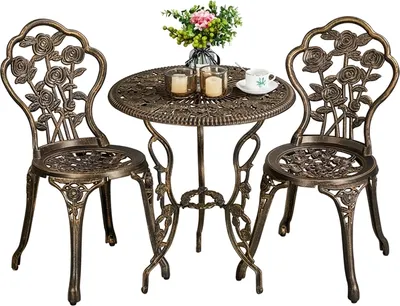 A vintage-style bronze bistro set with a round table and two chairs, decorated with rose motifs. A small vase of flowers and two candles are on the table.