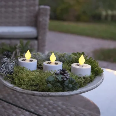 Three solar-powered LED tea lights are arranged in a decorative moss and pinecone display on a table, with a wicker chair in the background.