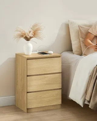 A bedside table with a vase of dried flowers and a phone charging next to a bed.