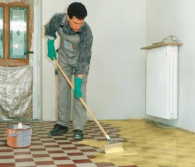 A man in overalls and green gloves is applying a yellow liquid to a checkered floor with a mop. There is a paint can on the floor and a radiator on the wall.