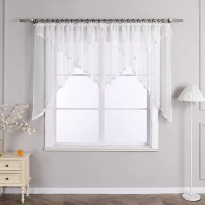 A white voile curtain with a scalloped edge and satin trim hangs in front of a window. A white floor lamp stands to the right, and a small cream-colored nightstand with two drawers is to the left.