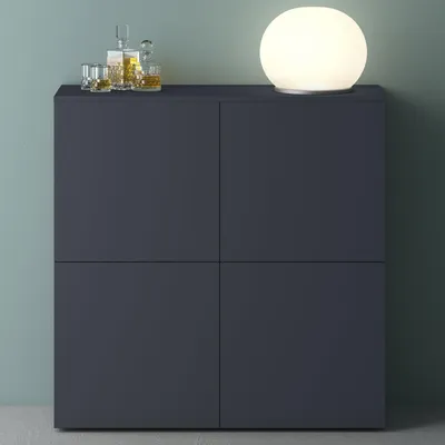 A dark gray cabinet with four doors, topped with two glasses of whiskey and a decanter, and a round white lamp on the right.