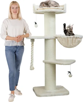 A woman stands next to a tall, cream-colored cat tree with multiple platforms and hanging toys. Two cats are visible on the cat tree.