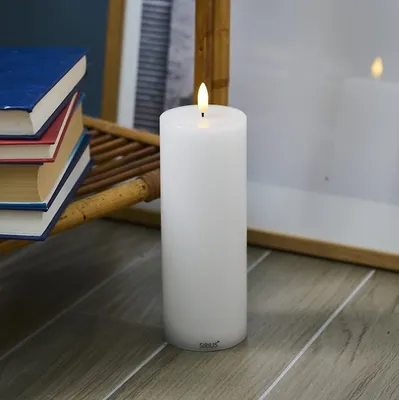 A white LED candle is placed on a wooden floor next to a stack of books and a mirror.