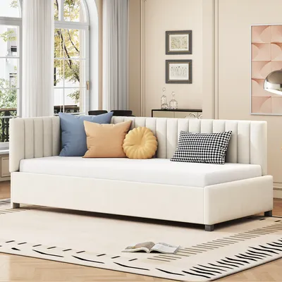A beige daybed with several decorative pillows is shown in a well-lit living room with large windows and sheer curtains.