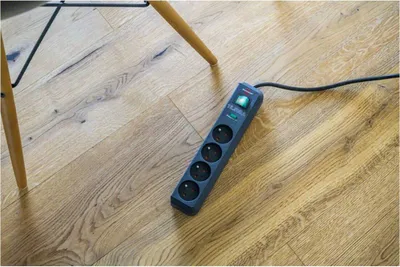 A black power strip with four outlets is lying on a wooden floor next to the legs of a chair.