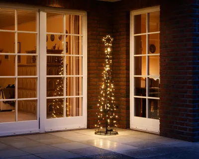A decorative LED Christmas tree with warm white lights stands on a patio next to a brick wall and large glass doors.