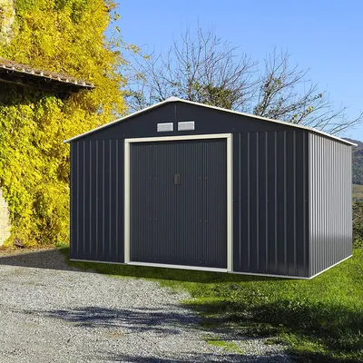 A dark gray metal shed with a double door is situated on a gravel driveway next to a building covered in yellow ivy, with trees and a blue sky in the background.