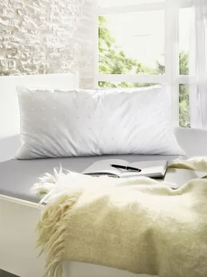 A pillow on a bed next to a window with a book and pen.