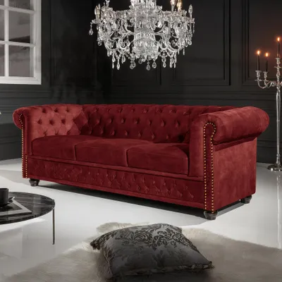 A luxurious red velvet Chesterfield sofa is displayed in a dimly lit room with a large crystal chandelier overhead and a candelabra in the background.
