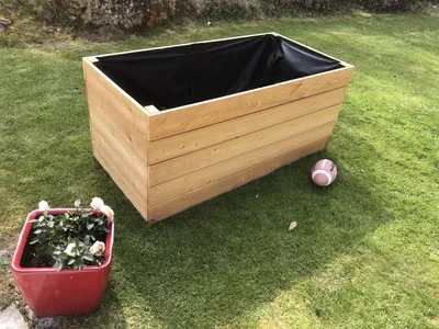 A wooden planter box is shown on a grassy lawn next to a red pot with a plant. A small ball is also on the grass.