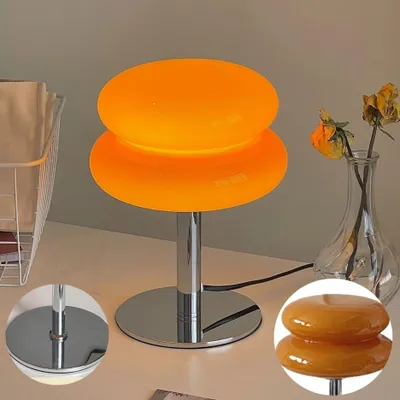 A modern orange mushroom-shaped table lamp sits on a desk next to a vase with dried flowers. A wire basket is visible on the left.