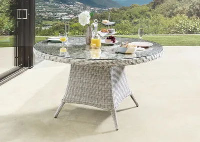 A round rattan dining table with a glass top, set on a patio with a scenic view of hills and buildings in the background. The table is set with drinks, fruit, and a tiered stand with snacks.