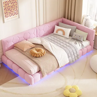 A pink corner bed with multiple pillows and blankets, decorated in a child's room with a rug and a picture on the wall.