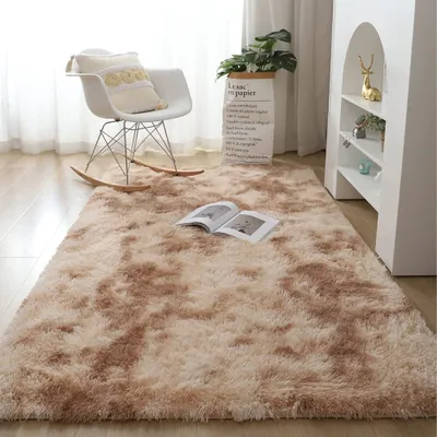 A fluffy beige rug with a tie-dye pattern is laid out on a wooden floor in a living room. A white rocking chair with a decorative pillow sits on the rug, and a paper bag with text is placed behind it. A white bookshelf with decorative items is visible on t
