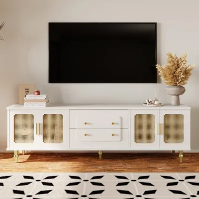 A white TV stand with cane doors and gold accents, featuring two drawers, is placed in a living room setting with a large black TV mounted on the wall above it. A rug with a black and white pattern is in the foreground, and decorative items like books and