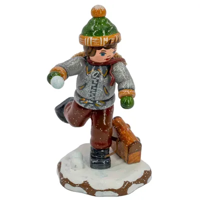 Ein kleiner Junge in Winterkleidung hält einen Schneeball in der Hand während er auf einem schneebedeckten Sockel steht und eine Schultasche neben ihm liegt