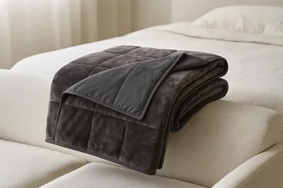 A weighted blanket is folded and draped over the armrest of a sofa, with a bed in the background.