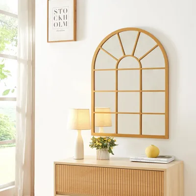 A gold arched mirror hangs above a wooden dresser with a lamp and a small vase of flowers.