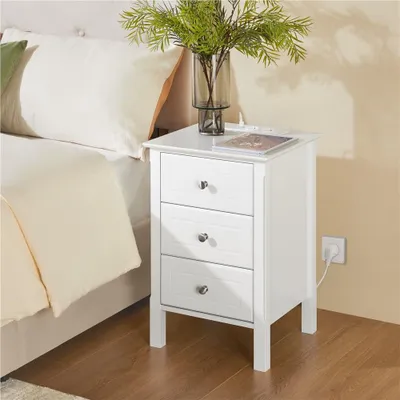 A white nightstand with three drawers is placed next to a bed. A vase with greenery sits on top of the nightstand.