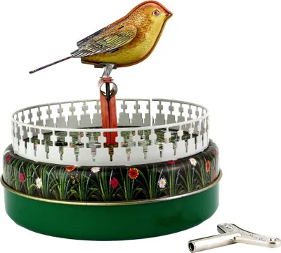 A vintage tin toy bird on a perch above a decorated cylindrical base, with a winding key in the foreground.