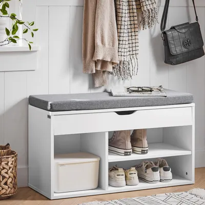 A white shoe bench with a gray cushion, featuring storage compartments for shoes and a drawer. It is placed against a white paneled wall, with clothes and a handbag hanging on hooks above.