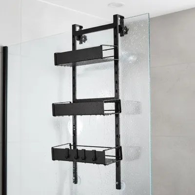 A black metal shower caddy with three baskets is hanging on a glass shower door.