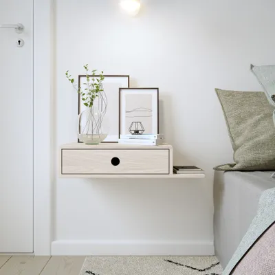 A floating nightstand with a drawer and a side shelf is mounted on a white wall next to a bed. The nightstand holds a vase with greenery and framed artwork. A phone is placed on the side shelf.
