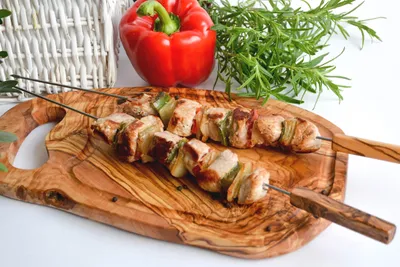 Two skewers of meat and vegetables are placed on a wooden cutting board, with a red bell pepper and rosemary sprigs in the background.
