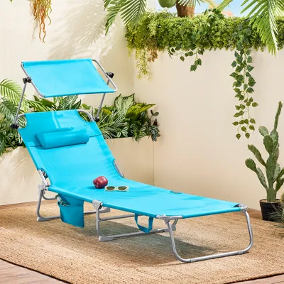 A blue chaise lounge with a canopy and pillow sits on a woven rug outdoors, surrounded by lush greenery and a cactus.