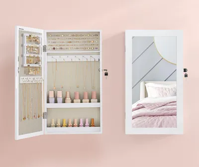 A white jewelry cabinet with its door open, displaying jewelry and nail polish, stands next to a full-length mirror reflecting a bedroom.