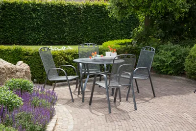A round garden table with four chairs set on a paved patio, surrounded by lush green hedges and purple flowers.