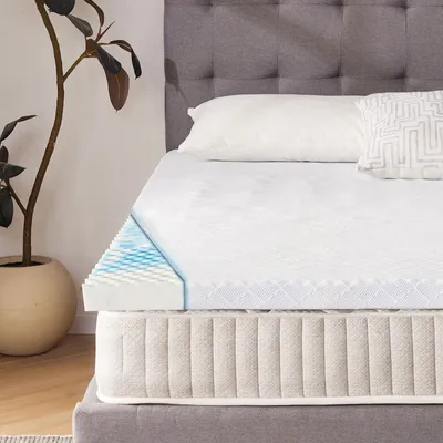 A mattress topper is shown on top of a mattress, with a cutaway view revealing its layers. A pillow and decorative pillow are on the bed, and a potted plant is to the left.