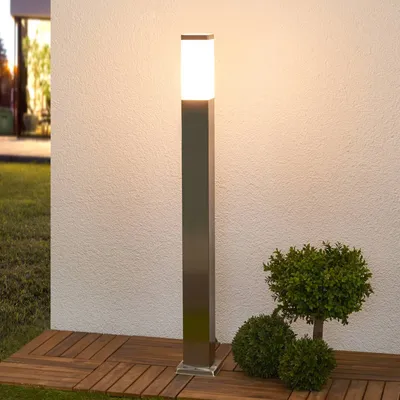 A modern outdoor lamp post with a square metal base and a frosted glass top, standing on a wooden deck next to some greenery.
