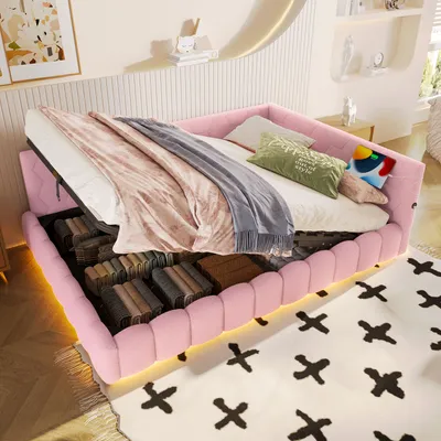 A pink upholstered bed with its storage compartment open, revealing folded items. The bed is made up with bedding and pillows, and is placed on a rug with a cross pattern. The room has a modern, minimalist aesthetic with decorative shelves and lighting.