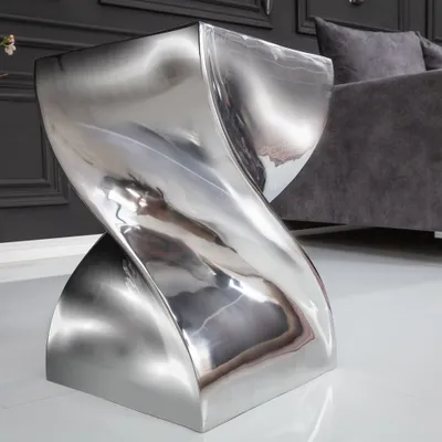 A highly polished silver side table or stool with a twisted design, placed on a white floor in a modern room with dark gray walls and a gray sofa in the background.