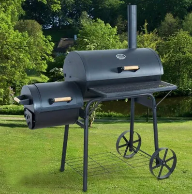 A large black barbecue grill with a side smoker attachment sits on a green lawn in front of trees and a body of water.