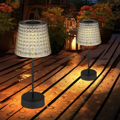 Two solar table lamps on a wooden deck at dusk, with plants and lanterns in the background.