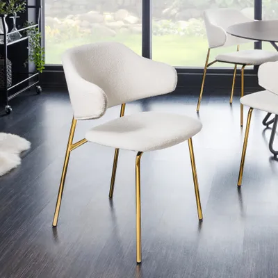 A white bouclé chair with gold metal legs is shown in a room with a dark floor and a large window in the background.