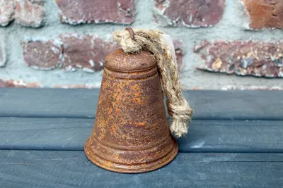 A rusty bell with a rope hangs in front of a brick wall and a wooden surface.
