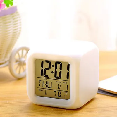 A white digital alarm clock cube is shown on a wooden surface, with a blurred background of a basket and greenery.