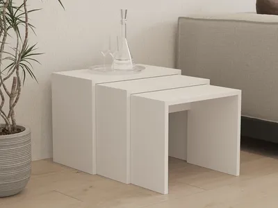 A set of three white nesting tables are arranged on a light wooden floor next to a potted plant and a sofa. Two wine glasses and a carafe are placed on the largest table.