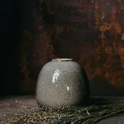 A speckled gray vase sits on a dark surface with dried grass in front of it, against a dark, textured background.